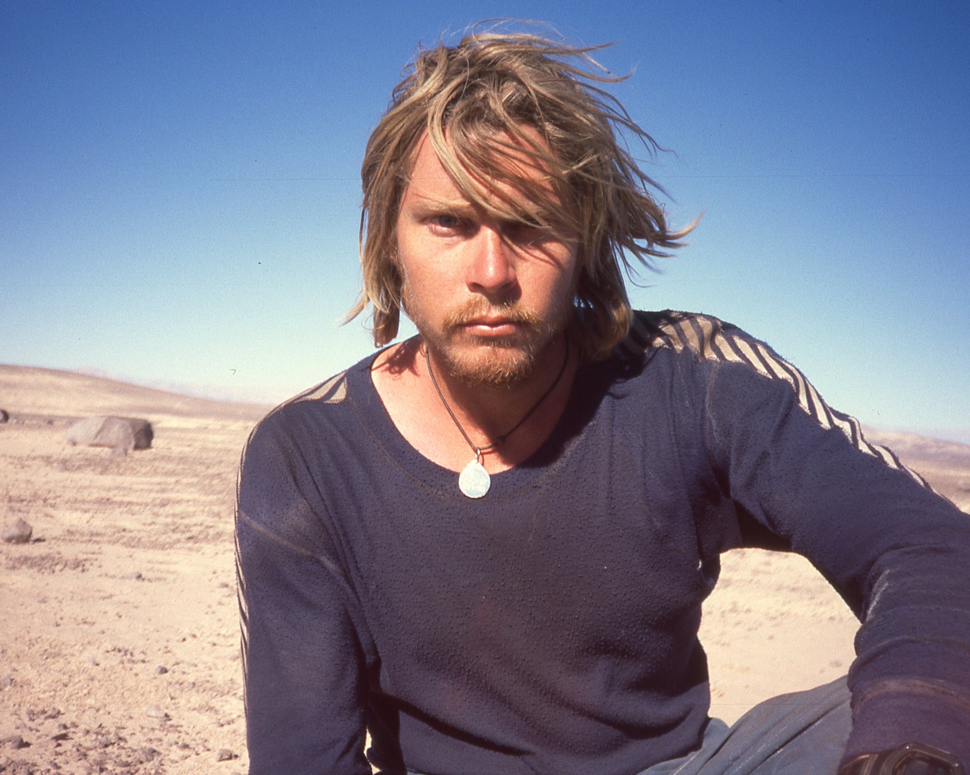 A younger Karl Bushby with long blond hair sits in a dry, sandy landscape in Chile in 1999.