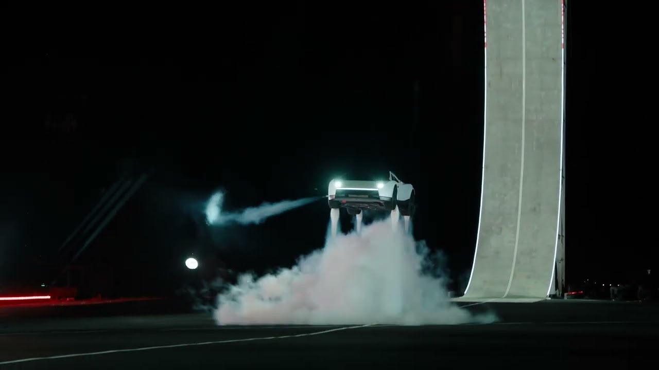 A white Tesla Roadster airborne beside a curved ramp, with gas plumes below it. Frame from Tesla’s The Ramp video, 15 October 2026.